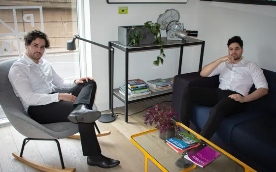 A photograph of Hussan sitting on a grey chair in the left corner and Hussain sitting on a sofa in the right corner of the photograph. Both are looking at the camera with relaxed postures.