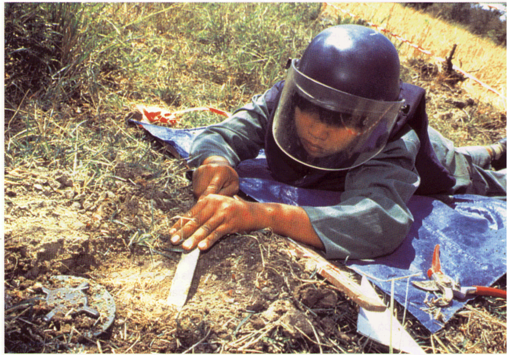A person lying on the ground in front of a landmine to try and demine it, wearing a protective face shield and vest.