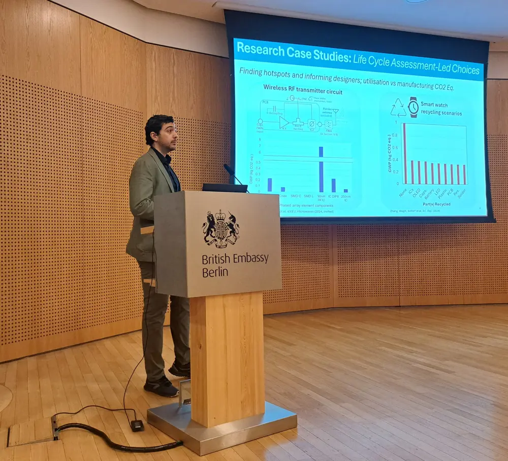 A man dressed smartly stands behind a lectern in a wood-panelled room with a big screen slightly behind him showing a few graphs