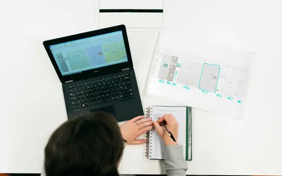An aerial view of an engineer writing in an A5 notebook, with a laptop and paper highlighted on the desk in front of them.