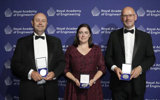 Two men stand either side of a woman, all wearing formal dress, in front of a blue background and all holding silver medals