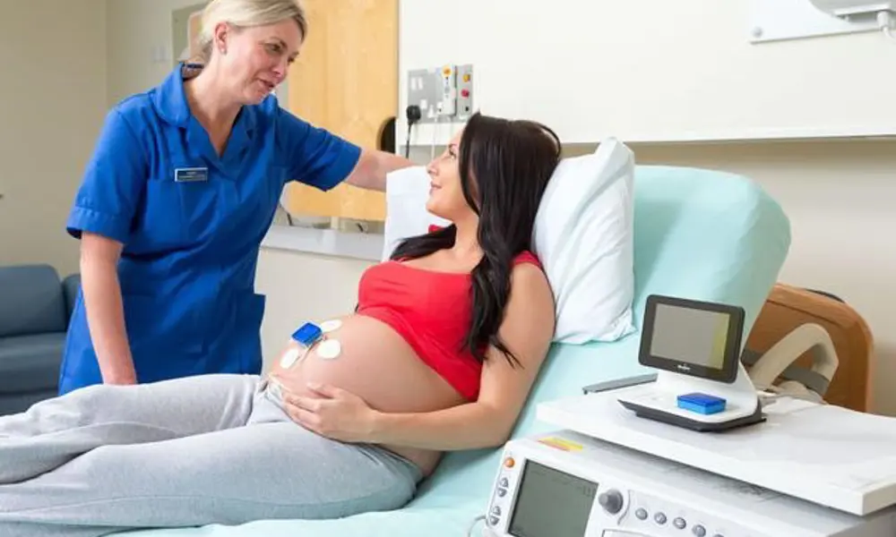 A pregnant women lying down on a hospital bed, talking to a nurse. The women is wearing the Monica Novii wireless patch system on her belly
