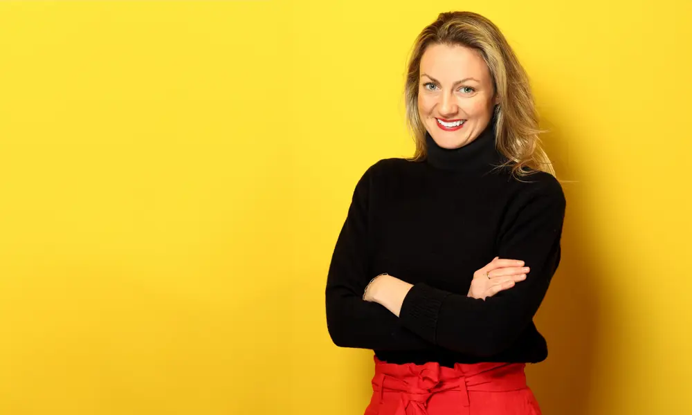 A headshot of Sinead O’Sullivan smiling at the camera, with her arms crossed and a yellow background.