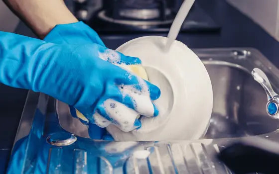Someone's hands wearing blue washing up gloves, cleaning a plate with a sponge and washing up liquid in the sink
