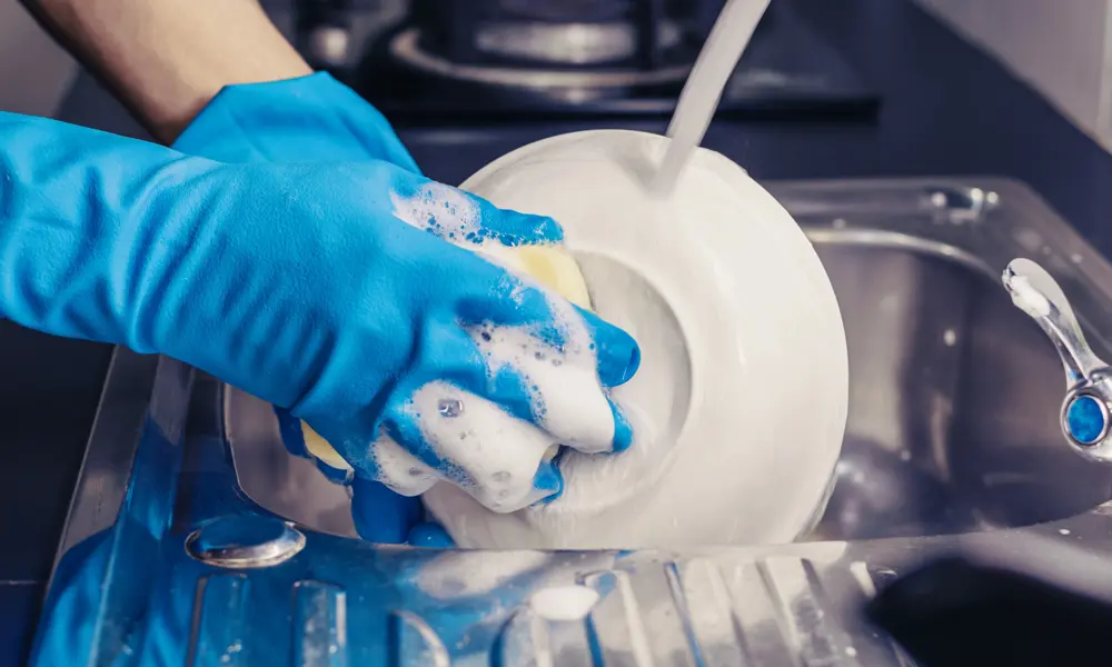 Someone's hands wearing blue washing up gloves, cleaning a plate with a sponge and washing up liquid in the sink