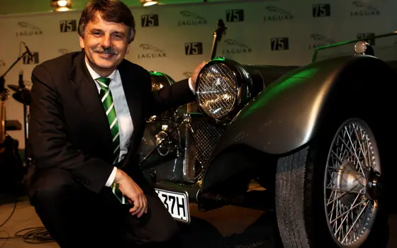 Ralf Speth, crouching next to an old Jaguar at a formal event in Germany.