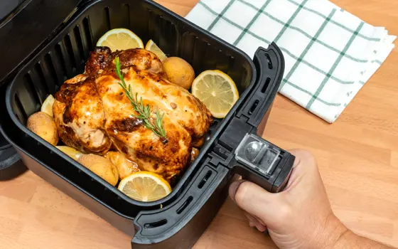 An air fryer basket, pulled out, holding a cooked roast chicken and sliced lemons 