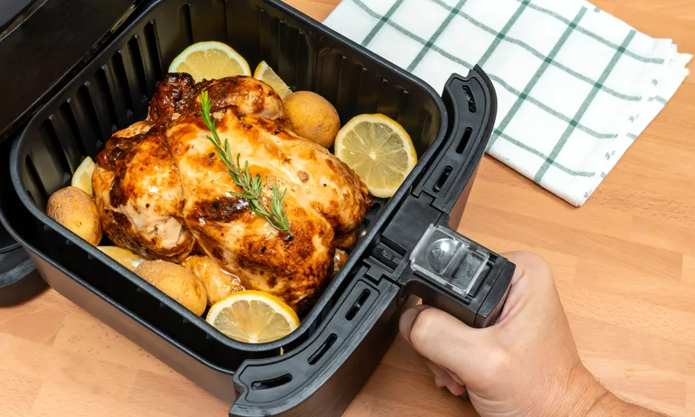 An air fryer basket, pulled out, holding a cooked roast chicken and sliced lemons 