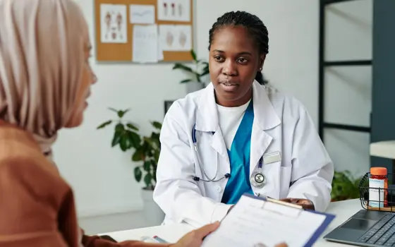 A Black female healthcare professional wearing a white medical jacket and stethoscope talks to a female patient wearing a peach hijab, who is holding a clipboard and form who  