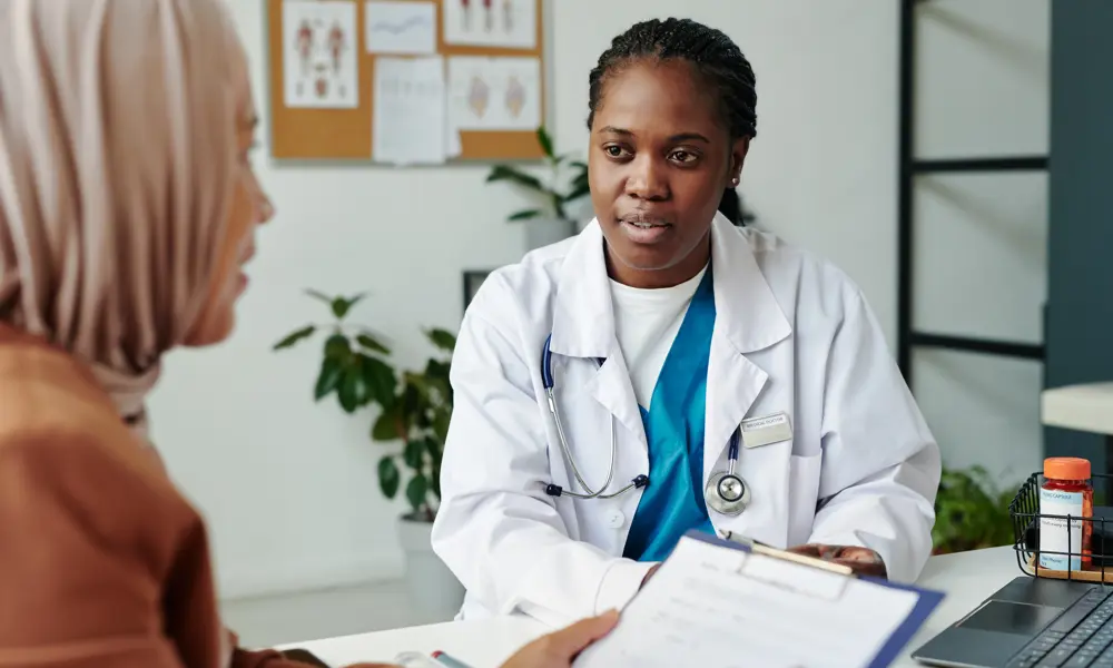 A Black female healthcare professional wearing a white medical jacket and stethoscope talks to a female patient wearing a peach hijab, who is holding a clipboard and form who  