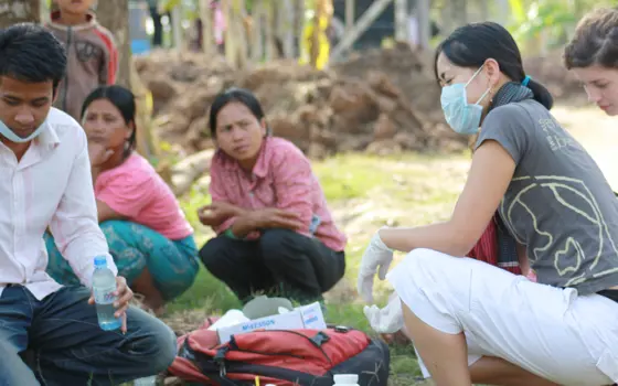 Dr Anh Tran crouching down with a mask and gloves on, with chemical bottles in front of her on the ground, being watched by local people in Cambodia.
