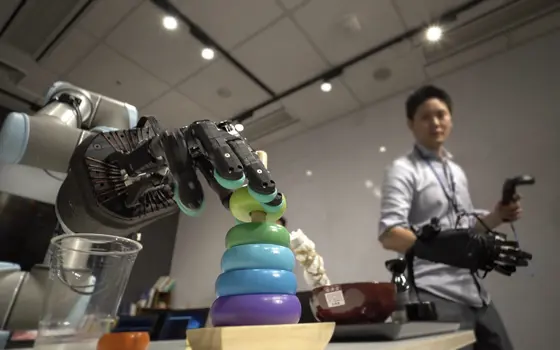 The Shadow Hand robot arm sliding a ring off a wooden stick. The hand is being controlled by a person in the background wearing a large glove. 