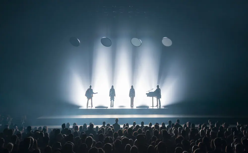 Two singers and two musicians playing a guitar and keyboard are brightly backlit by spotlights on a stage in front of a large crowd. 