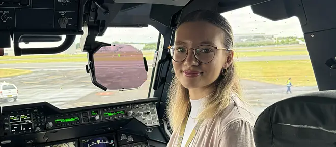 A blond woman wearing glasses sits inside a cockpit, looking at the camera