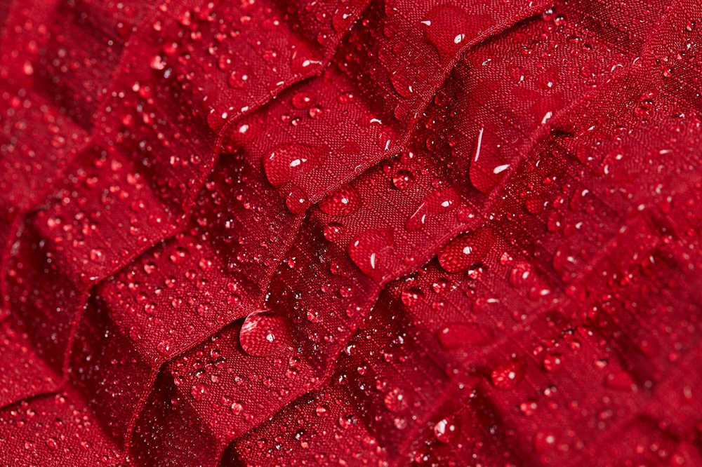A close-up of the red roof tile-like pattern that is used in petit pli's clothing with droplets of water on top of the fabric that demonstrate water resistance.