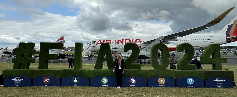 A woman stands outside in front of large green letters spelling #FIA2024 and a white aeroplane that has Air India on the side in red writing 