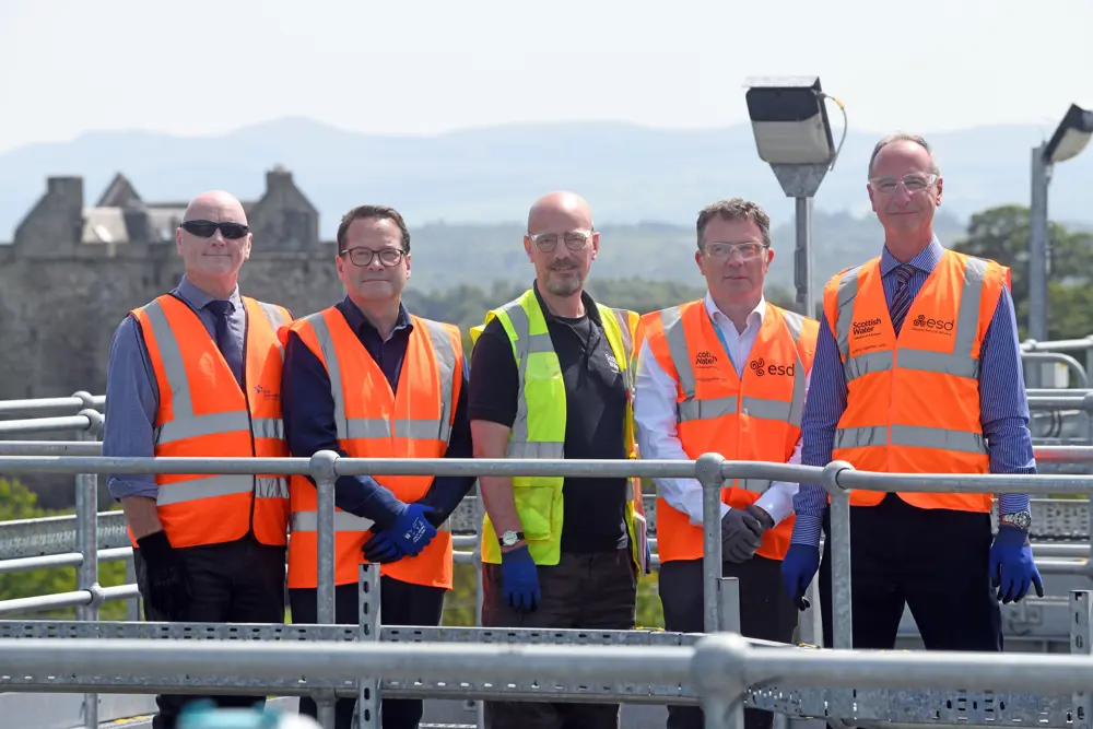 Five men, all wearing glasses and high vis vests, stand outside on top of a platform with grey handrails, with hills in the background
