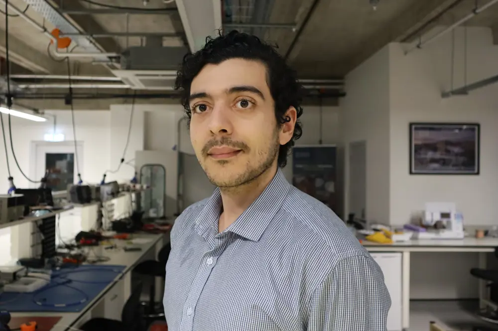 A man with brown hair wearing a blue shirt in an electronics lab looks beyond the camera