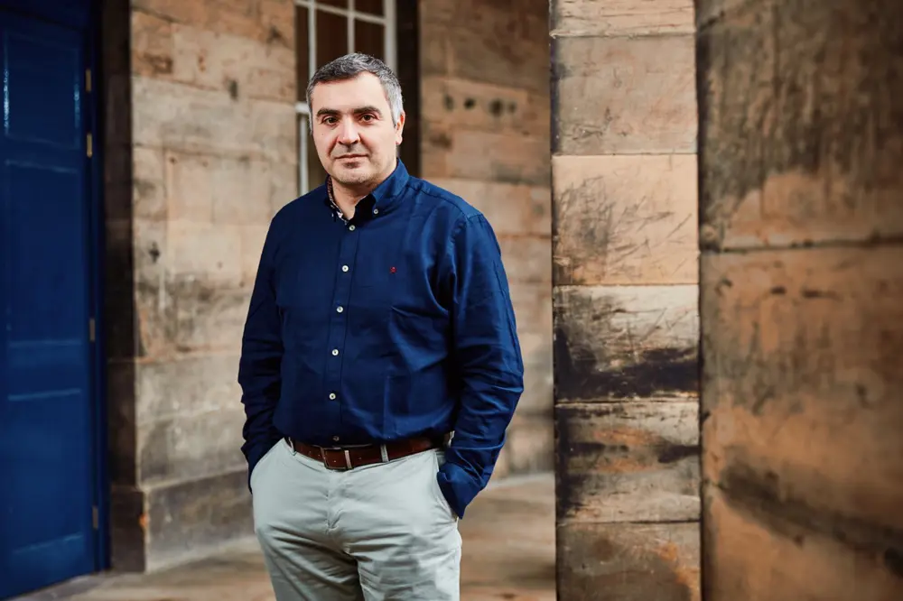 A portrait of Themis Prodromakis wearing a navy blue shirt and chinos, standing outside a historic building at the University of Edinburgh,