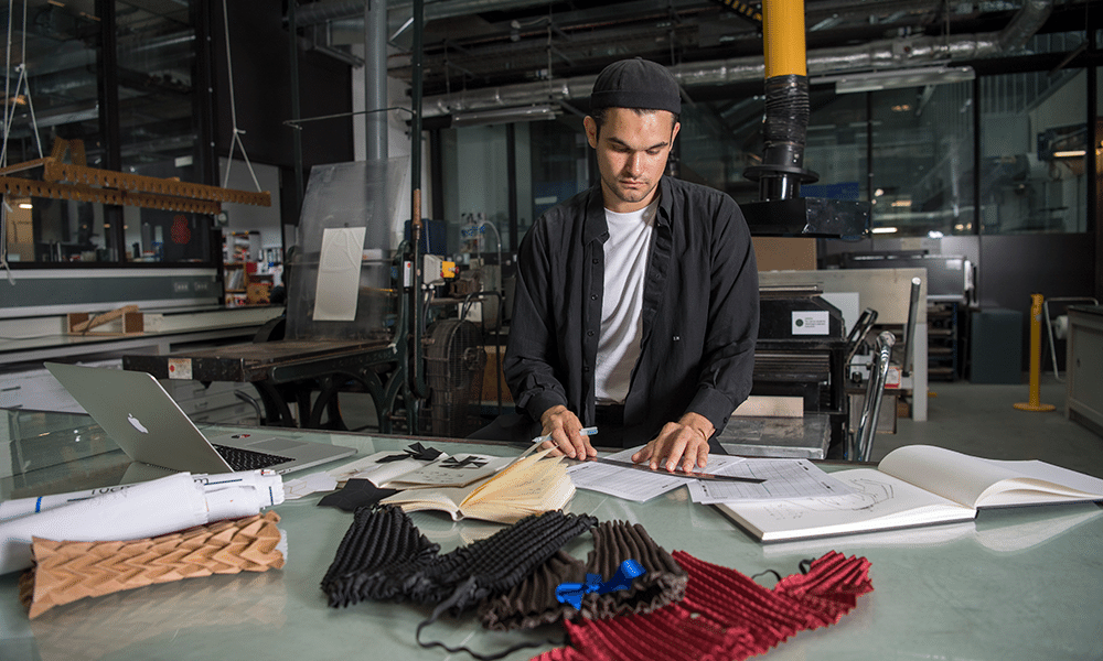 Ryan Mario Yasin in the studio behind a desk. He holds a ruler over his clothing designs and clothing samples can be seen on the desk in front of him. 