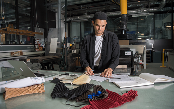 Ryan Mario Yasin in the studio behind a desk. He holds a ruler over his clothing designs and clothing samples can be seen on the desk in front of him.