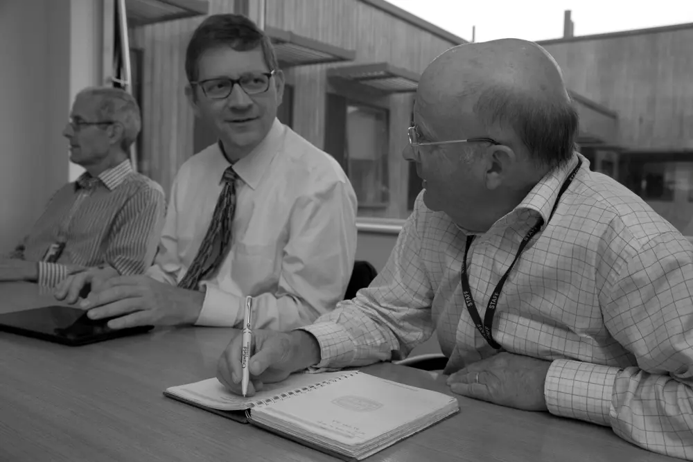 Black and white photograph of three men in business attire, sitting at a table in a meeting room with a pen and notebook.