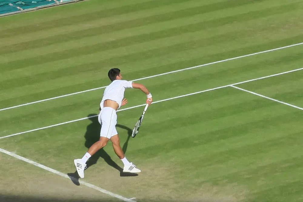 A man dressed in tennis whites and holding a tennis racket in motion on a grass tennis court 