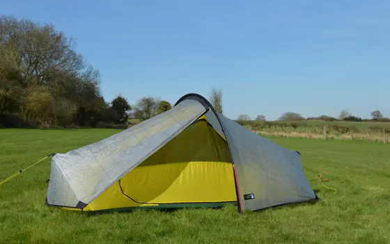 A terra nova tent in a field, with a grey outside and yellow inside lining. It is held up by a semi-circular polar in the middle, and vertical pole at each end, with pegs to keep the fabric in place.