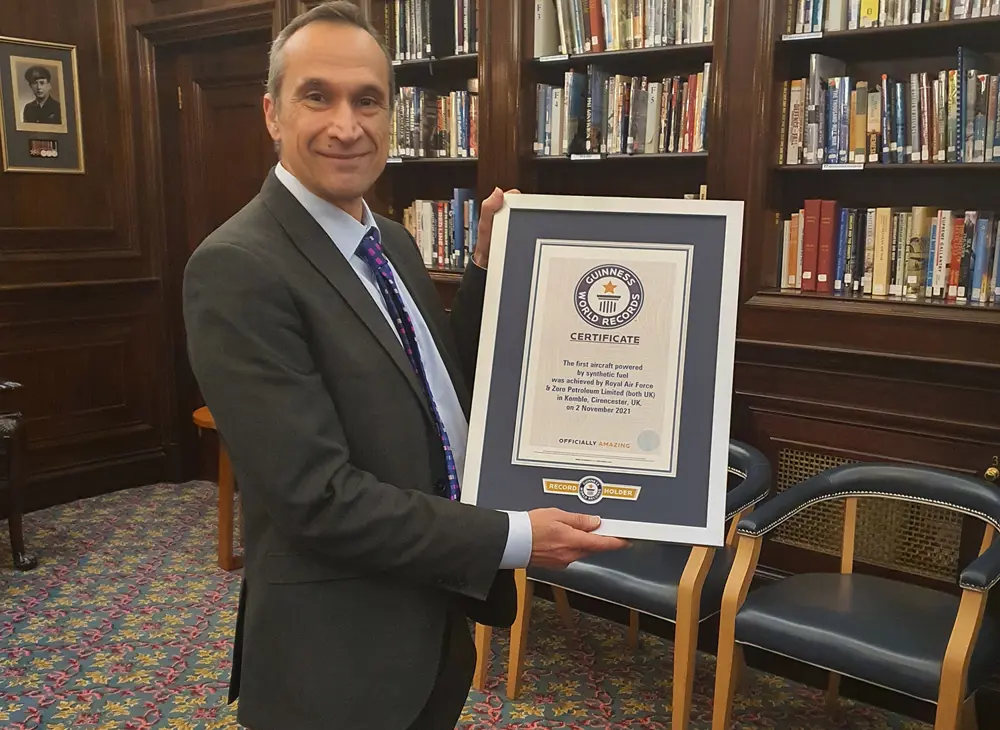 A man in a suit stands in a library smiling at the camera and holding up a certificate