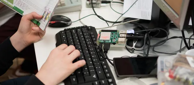 A child holding a book and typing on a keyboard, with a Raspberry Pi connected to the computer in front of them.