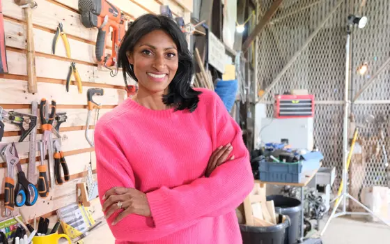 A woman wearing a bright pink jumper standing in a workshop with tools hanging on the wall