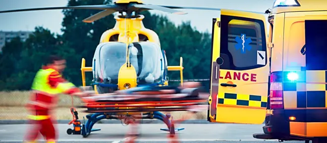 Paramedic in a red and neon yellow uniform transporting a patient from a helicopter into an ambulance.