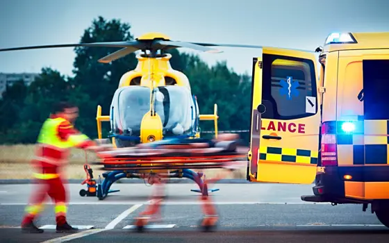 Paramedic in a red and neon yellow uniform transporting a patient from a helicopter into an ambulance.
