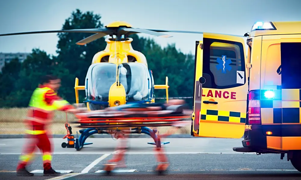 Paramedic in a red and neon yellow uniform transporting a patient from a helicopter into an ambulance.