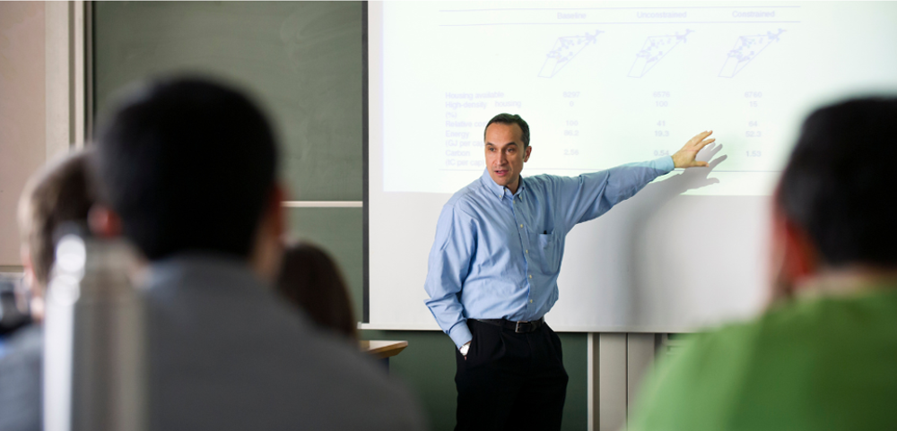 A man in a blue shirt gestures to a screen behind him in front of a room of people