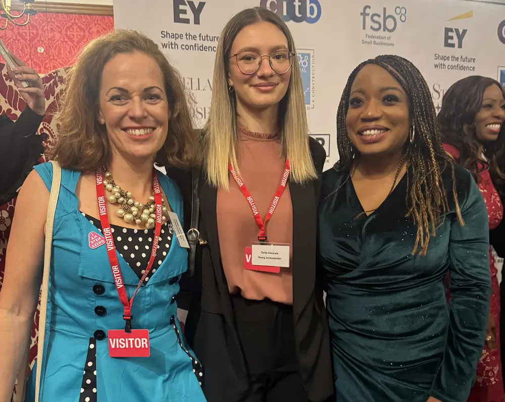 Three women in smart clothing pose in front of a backdrop, smiling at the camera