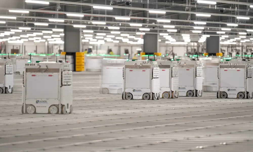 A well lit large room at the Erith warehouse, containing cube-shaped robots with wheels, on a large grid as a floor. 