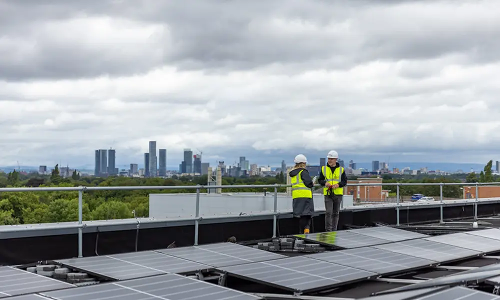 Two engineers wearing hi-vis jackets and hard hats standing talking on a solar panelled roof with a city skyline in the background