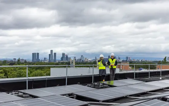 Two engineers wearing hi-vis jackets and hard hats standing talking on a solar panelled roof with a city skyline in the background