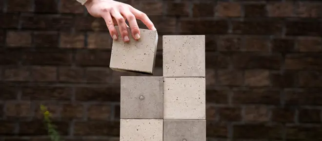 A hand places a small concrete block onto a stack of five others.