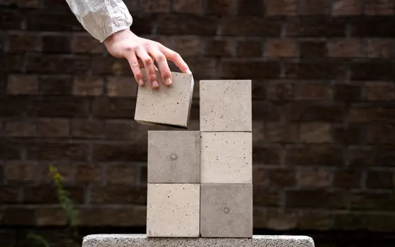 A hand places a small concrete block onto a stack of five others.