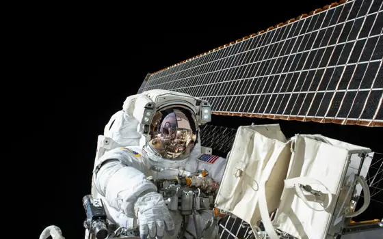 A astronaut spacewalking at the International Space Station, seen against the solar panels and the blackness of space.