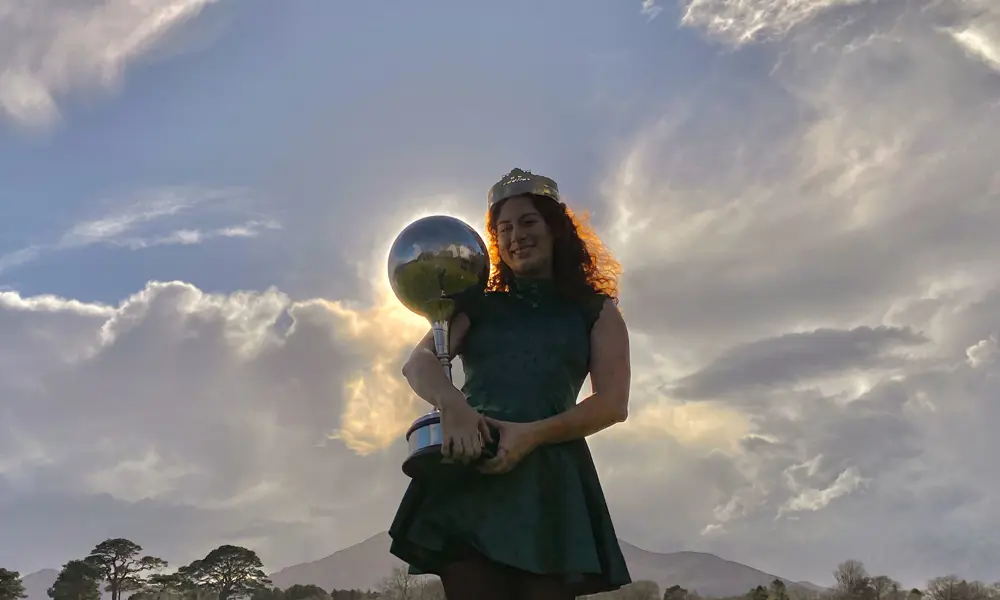 A woman with curly hear, wearing a crown and holding a trophy topped with a large sphere, that is positioned in front of the sun so there is the halo of light around it.