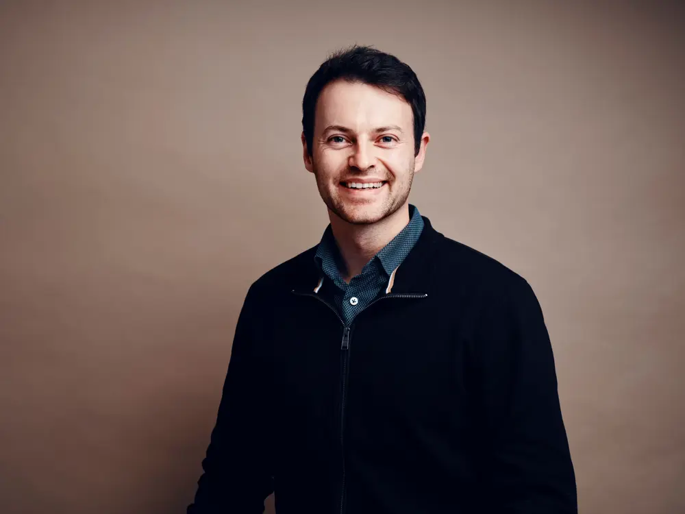 Alex Kendall smiling in a portrait photo, wearing a blue shirt and black zip up jacket.