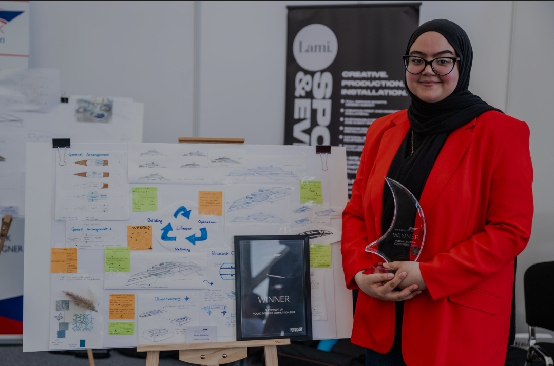 A woman wearing a red jacket, glasses and a headscarf holds a trophy in front of an easel