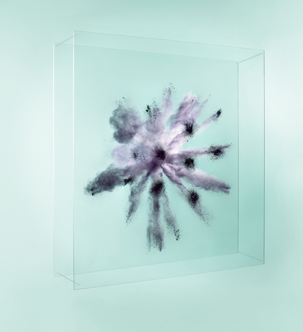 Black-grey powder explodes between two acrylic, transparent sheets, against a light turquoise background