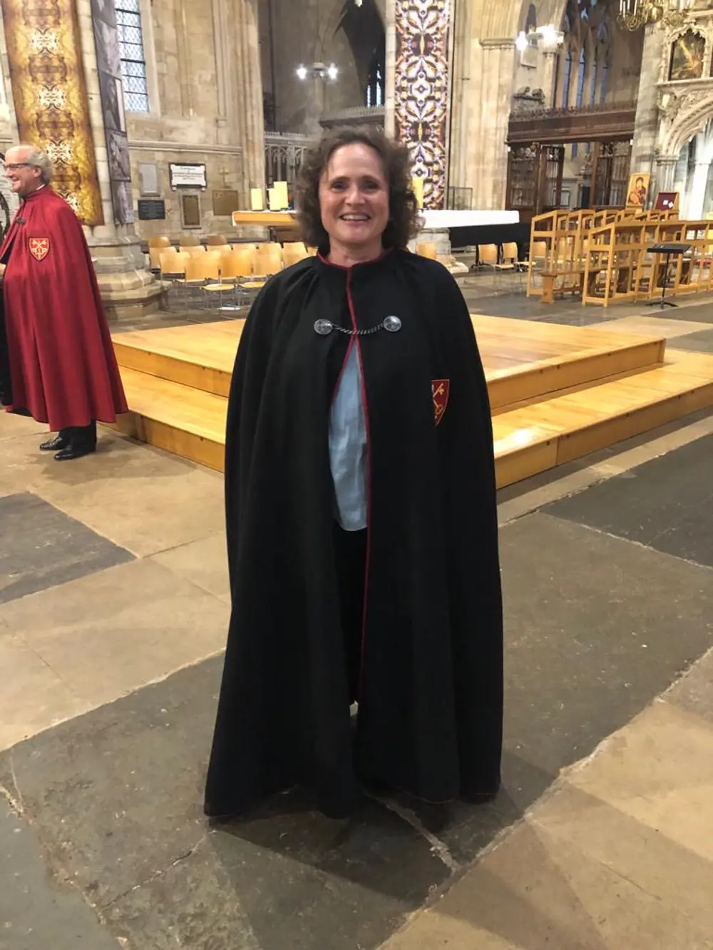 Penny has short dark hair, she is smiling in a cathedral, wearing a black robe.