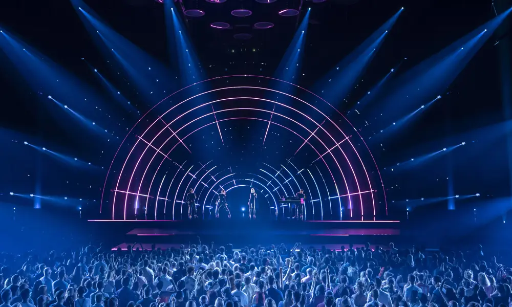 Two singers, a guitarist and a keyboard player play onstage to a large crowd, backlit by an LED lighting display that looks like a rainbow