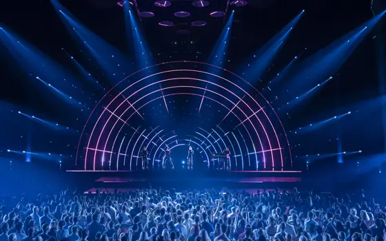 Two singers, a guitarist and a keyboard player play onstage to a large crowd, backlit by an LED lighting display that looks like a rainbow