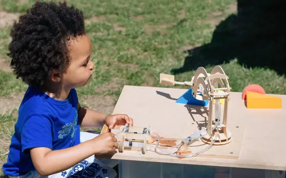 A young child playing outside with a wooden model containing syringes connected to plastic tubes. 